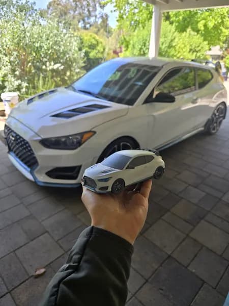 A custom scale model of a Hyundai Veloster N held up against the real car in a driveway