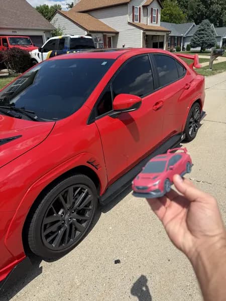 A hand holding a full-colour 3D printed Subaru WRX replica beside the owner's real car
