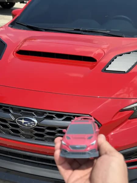 A miniature Subaru WRX replica held against the grille of the real car it was modelled from