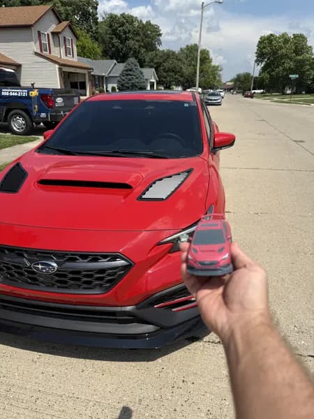 A 3D printed scale replica of a red Subaru WRX held in one hand in front of the real car