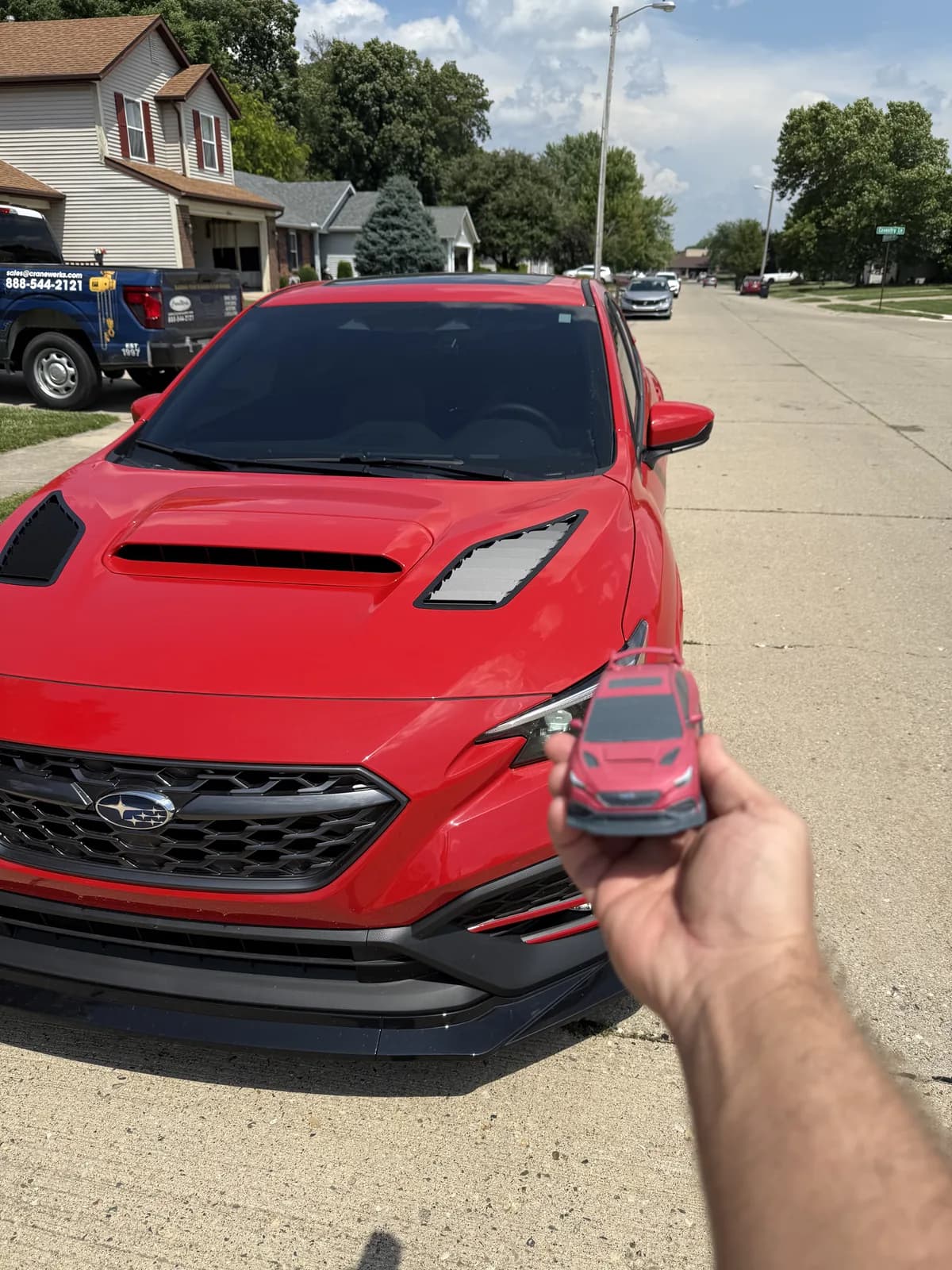 A 3D printed scale replica of a red Subaru WRX held in one hand in front of the real car